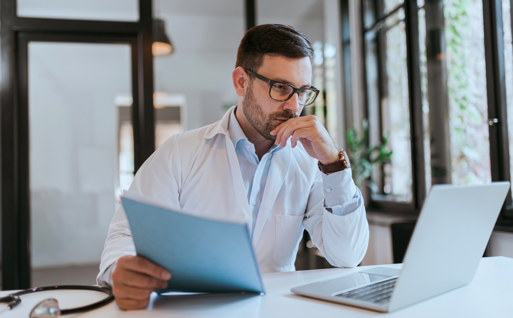Arzt an Schreibtisch vor Laptop mit Akte in der Hand