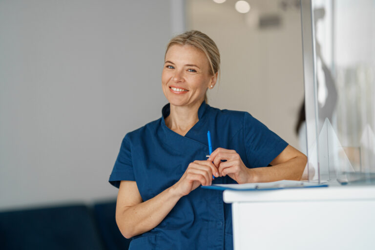 Smiling doctor with clipboard standing near reception in clinic hall and looking camera